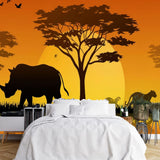 Papier Peint Animaux De La Savane dans une chambre à coucher apaisante avec lit blanc, table de chevet moderne, et plantes vertes. Illustration d'un coucher de soleil en savane africaine, avec des silhouettes de rhinocéros et de lions marchant à travers des herbes hautes, des arbres acacia en arrière-plan et des oiseaux volant dans le ciel.