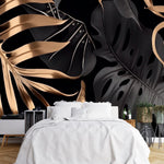 Papier Peint Panoramique Jungle Doré dans une chambre à coucher apaisante avec lit blanc, table de chevet moderne, et plantes vertes. Illustration de grandes feuilles de plantes tropicales noires et dorées sur un fond noir, avec des détails et textures métalliques, style moderne et élégant. 