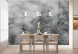 Papier Peint Noir Et Blanc Motif Vegetal dans une salle à manger contemporaine avec table en bois naturel, chaises beiges et éclairage suspendu blanc. Illustration d'une jungle dense et brumeuse avec des plantes tropicales luxuriantes et des oiseaux cachés parmi les branches, réalisée dans une palette de gris et verts foncés.