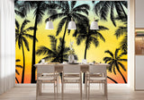 Papier Peint Panoramique Jungle Action dans une salle à manger contemporaine avec table en bois naturel, chaises beiges et éclairage suspendu blanc. Illustration d'une scène tropicale au coucher du soleil avec des silhouettes de palmiers en noir, sur un fond dégradé de bleu, jaune, orange et rouge.