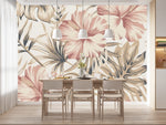 Papier Peint Imprimé Tropical Exotique dans une salle à manger contemporaine avec table en bois naturel, chaises beiges et éclairage suspendu blanc. Illustration d'un motif sophistiqué de feuilles tropicales et de fleurs d'hibiscus avec de grandes feuilles superposées et des détails minutieux, utilisant une palette de couleurs pastel composée de roses doux, crèmes et beige.