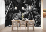 Papier Peint Panoramique Tropical Noir Et Blanc Exotique dans une salle à manger contemporaine avec table en bois naturel, chaises beiges et éclairage suspendu blanc. Illustration d'une vue de contre-plongée de grands palmiers illuminés par une lumière douce avec un ciel nocturne en arrière-plan, créant une ambiance apaisante et sereine.