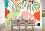 Papier Peint Jungle Panoramique Couleur dans une salle à manger contemporaine avec table en bois naturel, chaises beiges et éclairage suspendu blanc. Illustration d'art abstrait avec des motifs de feuilles tropicales dans différentes formes et couleurs, y compris vert, orange, rose, bleu marine, lavande, et blanc cassé, sur un fond blanc.