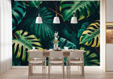 Tapisserie Adhesive Jungle dans une salle à manger contemporaine avec table en bois naturel, chaises beiges et éclairage suspendu blanc. Illustration de grandes feuilles de monstera tropicales dans une forêt dense, présentant diverses tailles et trous, équilibrées par d'autres feuillages exotiques luxuriants dans une palette de verts foncés et de touches de jaune.