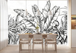 Papier Peint Vegetation Noir Et Blanc dans une salle à manger contemporaine avec table en bois naturel, chaises beiges et éclairage suspendu blanc. Illustration d'un ensemble dense de plantes tropicales, avec des éléments végétaux tels que des feuilles de bananier et des fougères, en dessin à l'encre noir sur fond blanc.