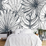 Papier Peint Feuille Tropical dans une chambre à coucher apaisante avec lit blanc, table de chevet moderne, et plantes vertes. Illustration d'un motif détaillé de feuilles tropicales avec de grandes feuilles superposées et un travail de lignes minutieux, réalisée dans un style noir et blanc.