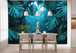 Papier Peint Panoramique Jungle Bleu dans une salle à manger contemporaine avec table en bois naturel, chaises beiges et éclairage suspendu blanc. Illustration d'une forêt tropicale dense avec des feuilles de monstera luxuriantes et autres végétations, baignant dans une lumière douce et mystique.