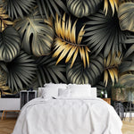 Papier Peint Tropical Noir Doré dans une chambre à coucher apaisante avec lit blanc, table de chevet moderne, et plantes vertes. Illustration d'un motif luxueux de feuilles tropicales avec de grandes feuilles superposées et des détails minutieux, utilisant des couleurs noir et or avec des textures contrastantes sur un fond sombre.