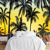 Papier Peint Panoramique Jungle Action dans une chambre à coucher apaisante avec lit blanc, table de chevet moderne, et plantes vertes. Illustration d'une scène tropicale au coucher du soleil avec des silhouettes de palmiers en noir, sur un fond dégradé de bleu, jaune, orange et rouge.