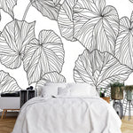 Papier Peint Motif Vegetal Noir Et Blanc dans une chambre à coucher apaisante avec lit blanc, table de chevet moderne, et plantes vertes. Illustration de feuilles tropicales abstraites avec des lignes ondulées en noir sur fond clair, dans un style minimaliste et contemporain.