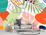 Papier Peint Jungle Panoramique Couleur dans une chambre de bébé moderne et minimaliste avec des murs blancs et un sol foncé, un berceau blanc, une couverture grise à pompons, des peluches, des jouets en bois et un tapis à motif noir et blanc. Illustration d'art abstrait avec des motifs de feuilles tropicales dans différentes formes et couleurs, y compris vert, orange, rose, bleu marine, lavande, et blanc cassé, sur un fond blanc.