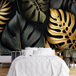 Papier Peint Tropical Noir dans une chambre à coucher apaisante avec lit blanc, table de chevet moderne, et plantes vertes. Illustration d'un motif luxueux de feuilles tropicales avec de grandes feuilles superposées et des détails minutieux, utilisant des couleurs noir et or avec des textures contrastantes sur un fond sombre.