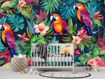 Papier Peint Jungle Perroquet Tropical dans une chambre de bébé moderne et minimaliste avec des murs blancs et un sol foncé, un berceau blanc, une couverture grise à pompons, des peluches, des jouets en bois et un tapis à motif noir et blanc. Illustration de trois perroquets colorés perchés sur des branches entourées de feuillage tropical luxuriant et de fleurs colorées.