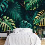 Tapisserie Adhesive Jungle dans une chambre à coucher apaisante avec lit blanc, table de chevet moderne, et plantes vertes. Illustration de grandes feuilles de monstera tropicales dans une forêt dense, présentant diverses tailles et trous, équilibrées par d'autres feuillages exotiques luxuriants dans une palette de verts foncés et de touches de jaune.