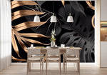 Papier Peint Panoramique Jungle Doré dans une salle à manger contemporaine avec table en bois naturel, chaises beiges et éclairage suspendu blanc. Illustration de grandes feuilles de plantes tropicales noires et dorées sur un fond noir, avec des détails et textures métalliques, style moderne et élégant. 