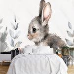 Papier Peint Enfant Animaux dans une chambre à coucher apaisante avec lit blanc, table de chevet moderne, et plantes vertes. Illustration d'un lapin gris assis dans un champ, entouré de plantes colorées dans un style aquarelle détaillé.