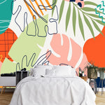 Papier Peint Jungle Panoramique Couleur dans une chambre à coucher apaisante avec lit blanc, table de chevet moderne, et plantes vertes. Illustration d'art abstrait avec des motifs de feuilles tropicales dans différentes formes et couleurs, y compris vert, orange, rose, bleu marine, lavande, et blanc cassé, sur un fond blanc.