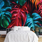 Papier Peint Tropical Coloré dans une chambre à coucher apaisante avec lit blanc, table de chevet moderne, et plantes vertes. Illustration d'un motif vibrant de feuilles tropicales avec de grandes feuilles superposées aux détails minutieux, utilisant des couleurs rouges profonds, verts riches et oranges éclatants.