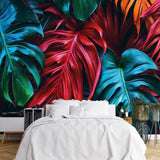 Papier Peint Tropical Coloré dans une chambre à coucher apaisante avec lit blanc, table de chevet moderne, et plantes vertes. Illustration d'un motif vibrant de feuilles tropicales avec de grandes feuilles superposées aux détails minutieux, utilisant des couleurs rouges profonds, verts riches et oranges éclatants.