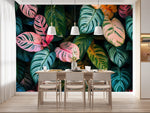 Papier Peint Feuillage Tropical Exotique dans une salle à manger contemporaine avec table en bois naturel, chaises beiges et éclairage suspendu blanc. Illustration d'un motif vibrant de feuilles tropicales avec de grandes feuilles superposées et des détails minutieux, utilisant des couleurs roses, verts et oranges avec des motifs et textures variés.