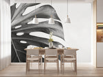 Tapisserie Noire Tropicale Salon dans une salle à manger contemporaine avec table en bois naturel, chaises beiges et éclairage suspendu blanc. Illustration en gros plan d'une feuille de monstera en noir et blanc, dans un style minimaliste et moderne.